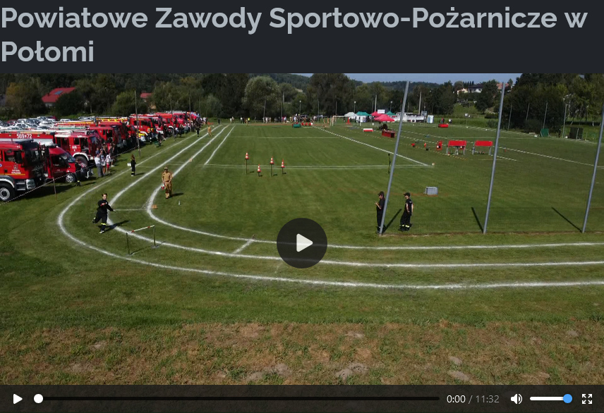 Kadr z nagrania wideo przedstawiający Powiatowe Zawody Sportowo-Pożarnicze w Potomii. W centrum widoczna jest zielona murawa boiska, na której wyznaczone są białe linie torów do biegu. Na torze biegnie strażak, prawdopodobnie biorący udział w zawodach. W tle po lewej stronie ustawione są rzędy czerwonych wozów strażackich, a po prawej stronie boiska znajdują się przeszkody i sprzęt wykorzystywany w zawodach. Wzdłuż toru stoją inni strażacy w ciemnych mundurach, obserwując przebieg rywalizacji. Na górze ekranu widnieje napis "Powiatowe Zawody Sportowo-Pożarnicze w Potomii".