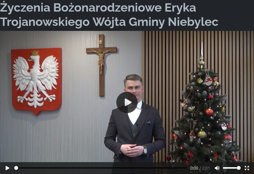 Wójt Gminy Niebylec stoi na tle choinki udekorowanej bombkami i światełkami oraz godła Polski i krzyża na ścianie. Na górze ekranu znajduje się napis: „Życzenia Bożonarodzeniowe Eryka Trojanowskiego Wójta Gminy Niebylec”. Na dole widoczny jest pasek odtwarzacza wideo z czasem trwania filmu: 2:01.