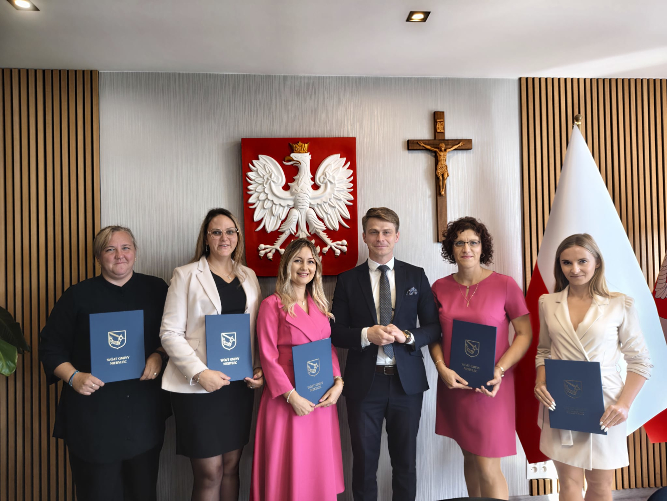 Pięć nauczycielek wraz z Wójtem Gminy Niebylec podczas uroczystego wręczenia aktów nadania stopnia nauczyciela mianowanego. Nauczycielki trzymają granatowe teczki z herbem Gminy Niebylec. W tle widoczne są godło Polski oraz flaga.