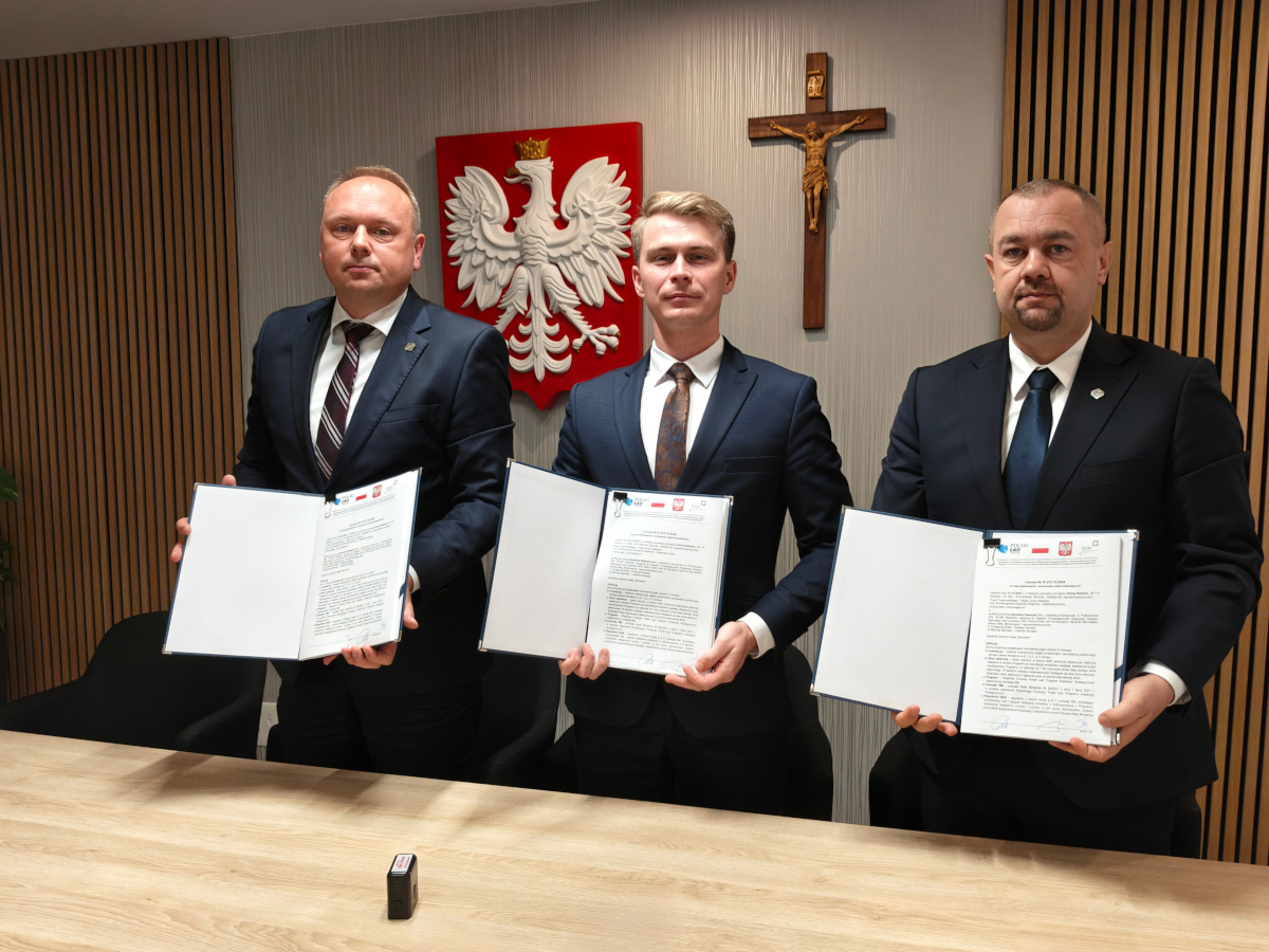 Trzech mężczyzn w garniturach stoi obok siebie, trzymając otwarte teczki z dokumentami, które pokazują w stronę aparatu. W tle widać godło Polski oraz krzyż. Pomieszczenie ma drewniane panele na ścianie po lewej stronie.